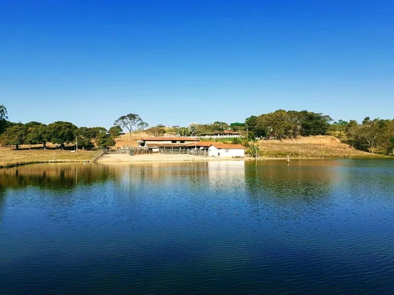 Lago de água escura refletindo o céu azul, com construções térreas e pasto na margem oposta