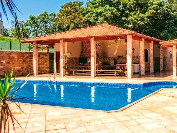 Piscina azul em piso de pedra clara, diante de um quiosque de telhado cerâmico com mesas de madeira